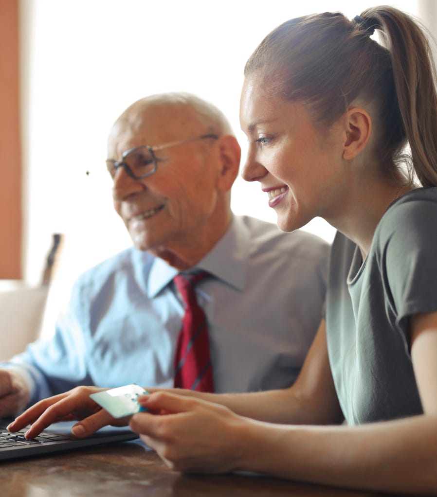 Young woman in casual clothes helping senior man in formal shirt with paying credit card in Internet using laptop while sitting at table